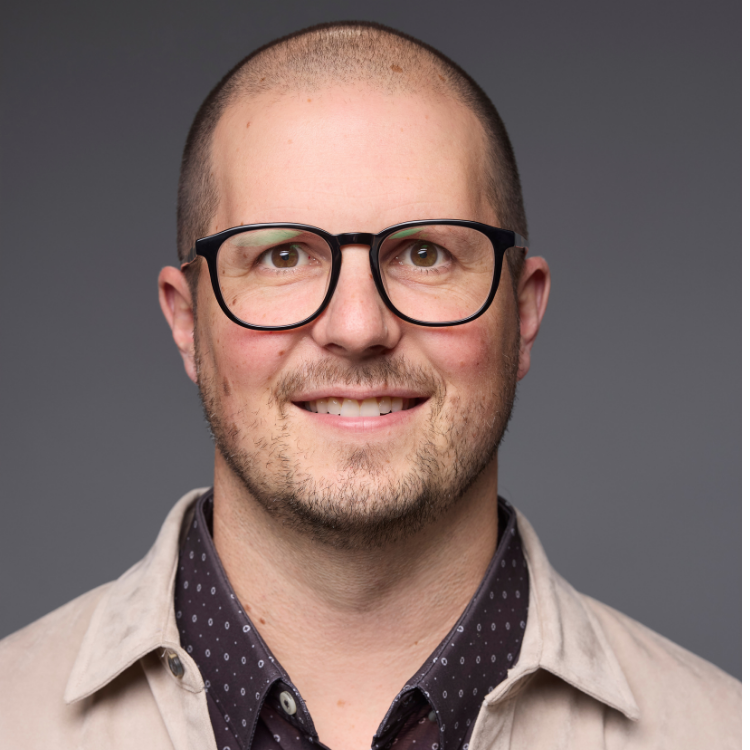 A smiling man with a shaved head, glasses, and light facial hair wears a beige jacket over a dark, patterned collared shirt. The background is plain and gray.