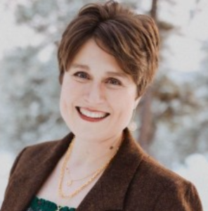 Jessa Crisp, a woman with short brown hair and a brown blazer, smiles outdoors. She wears layered gold necklaces and a green top. The background is blurred with trees and appears to be snowy.