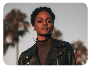 A woman wearing a black leather jacket and hoop earrings stands outdoors with palm trees in the background, softly lit by the setting sun.