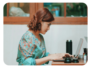 A woman with curly hair and a patterned teal top sits at a desk typing on a laptop, with a tumbler and a bottle nearby, in front of a window.
